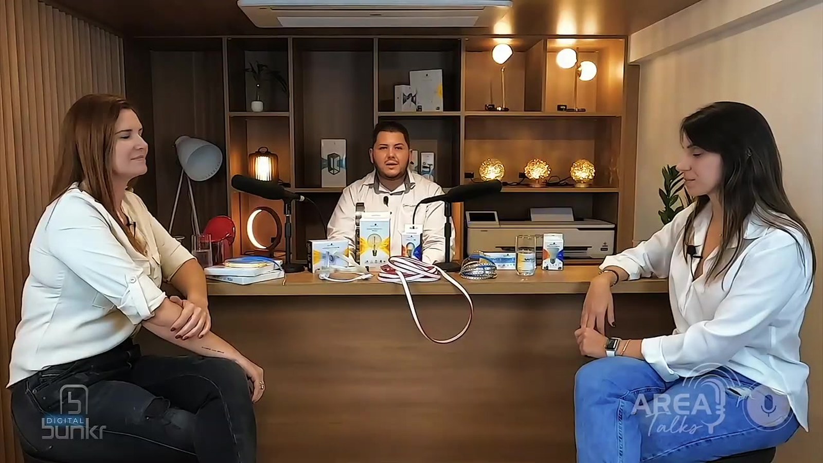 Quadro do programa Área Talks: apresentador atrás de uma bancada com microfones e produtos, entre duas convidadas sentadas, com o logo do programa no canto