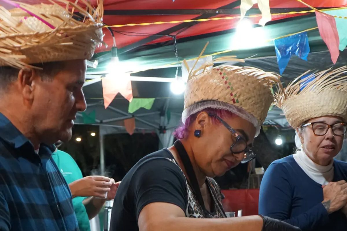 Três pessoas de chapéu de palha trabalhando atrás do balcão da barraca, à noite, sob as bandeirinhas