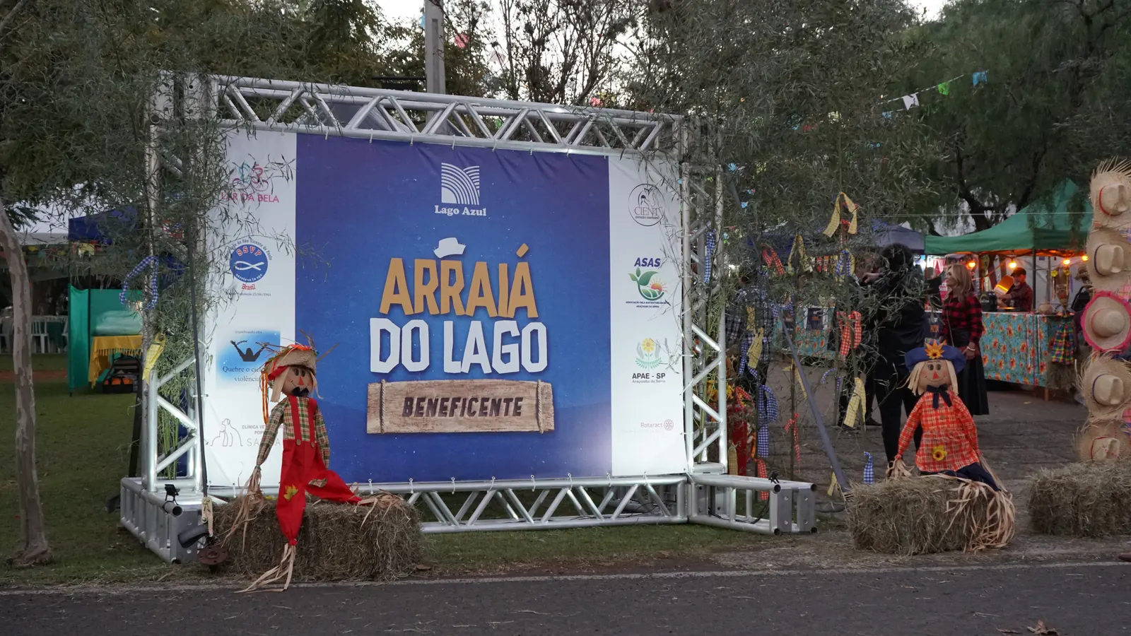 Painel impresso montado em treliça na entrada da festa, com a arte do Arraiá do Lago Beneficente ao centro e os logotipos das instituições nas duas laterais
