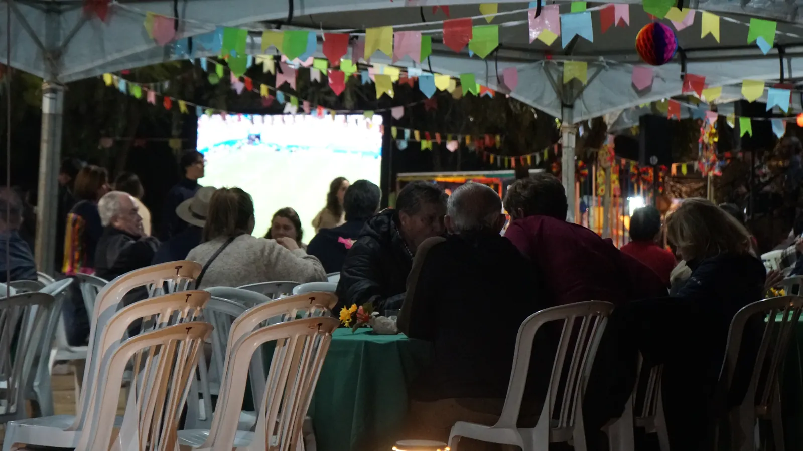 Mesas ocupadas sob a tenda decorada com bandeirinhas e balão de papel, com um telão aceso ao fundo