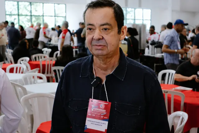 Participante com crachá do congresso e microfone de lapela preso à camisa, com o salão cheio e as mesas de toalha vermelha ao fundo