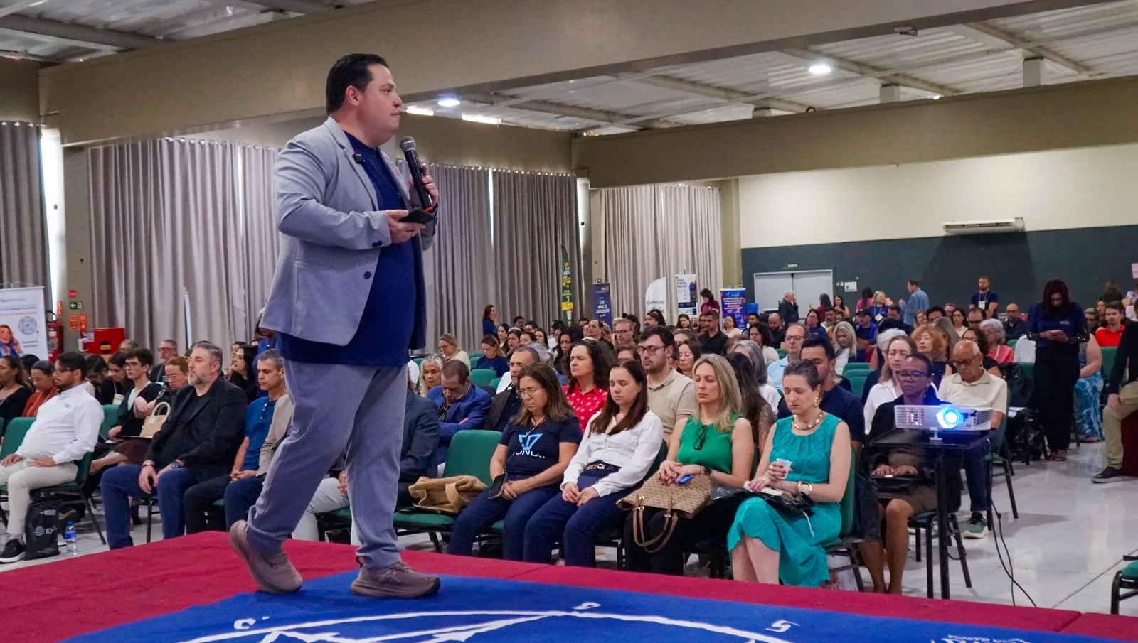 Palestrante de blazer cinza anda pelo palco de tapete azul com o microfone na mão, diante do salão cheio de participantes sentados